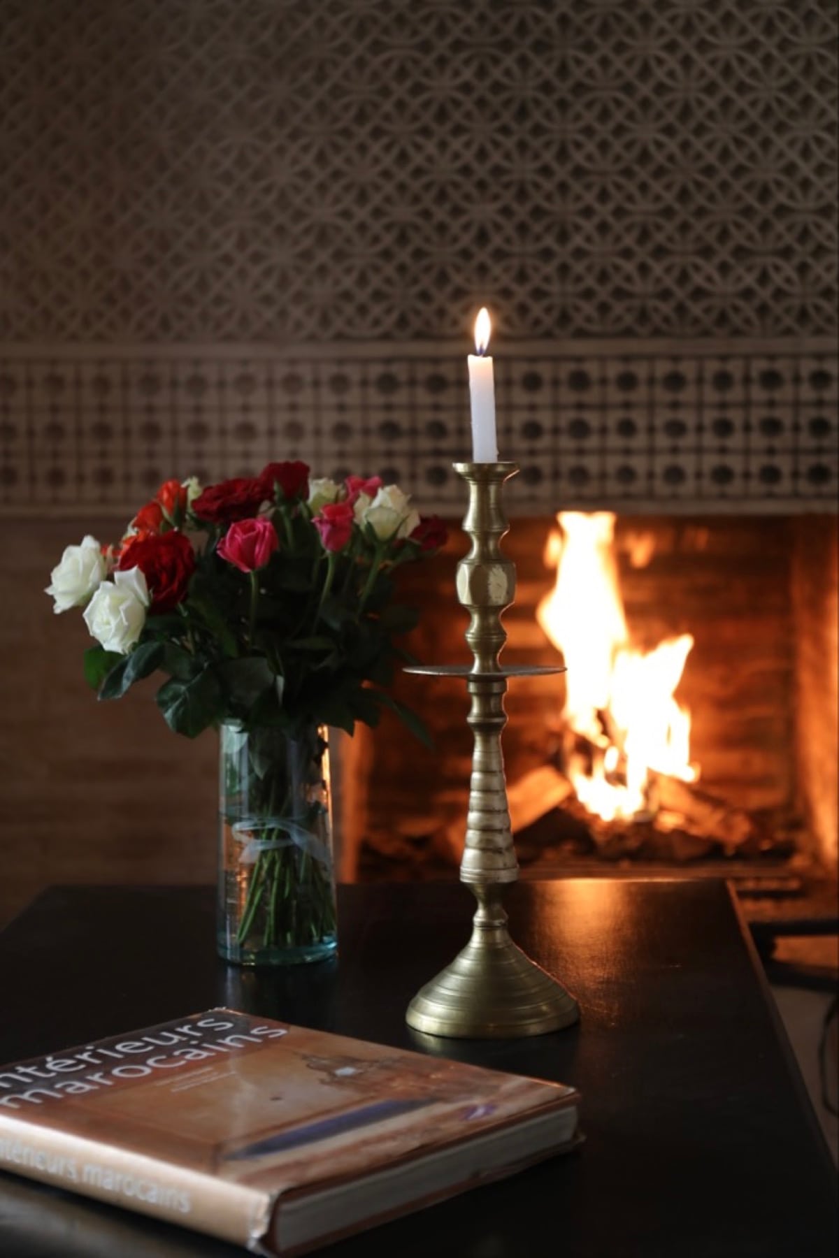A lit fireplace reflected beside flowers and a brass candlestick